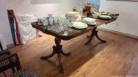Wide angle photo showing the wooden dining table with various dishes and glassware on top. Two brass-accented pedestal legs visible, wood surface has scratches.