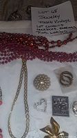 Photo of various bead necklaces in red, coral, white, and blue colors, assorted brooches including a pearl cluster and decorative pins, and metal earrings.