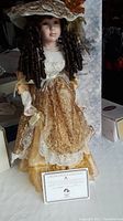 Porcelain doll Maggie dressed in golden lace Victorian-style gown with large hat, curly brown hair, pearl necklace, and Certificate of Authenticity visible.