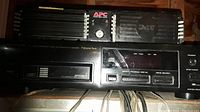 Photo of black electronic equipment units stacked, showing APC power conditioner on top with power switch, black audio receiver/amplifier below, and Optimus CD player with cassette deck below that. Cables visible at front below devices.