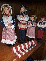 Close-up photo of four 4th of July themed caroler figurines and two rolled striped fabric decorations on wooden surface