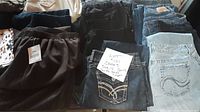 Photo showing several pairs of women's jeans in blue and black denim, black skirt, and cream dress folded on a surface with a note indicating size 14 and lot details.