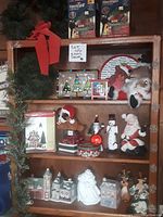 Full display of lot items on wooden bookshelf showing various Christmas decor including Dept 56 houses, Santa, snowman, wreath, tins, and plush Santa figure.