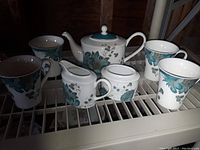Photo showing full 7-piece set on rack including teapot, 4 cups, sugar bowl, and creamer with teal floral pattern.