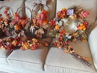 Photo showing a collection of artificial fall decor arranged on a couch, including wreath, floral baskets, and decorative stick bundles, highlighting seasonal colors and materials.