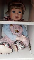Close-up of a porcelain doll dressed in pastel overalls with plaid trim and sitting in original box, detailed with painted blue eyes and realistic facial features.