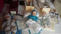 Three Yolanda Bello baby dolls displayed with original boxes, certificates, and musical blankets. Dolls wear soft fabric clothes and have porcelain heads, arms, and feet.