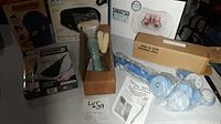 Photo showing boxed HoMedics Neck Massager, Spin Spa base and attachments still in plastic, Shiatsu Foot Massager box, and other massage brushes and gloves on table.