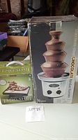 Photo showing the boxed stainless steel chocolate fondue fountain next to the boxed Feng Shui tranquility fountain on a table.