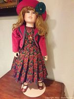 Full view of the Bette Ball porcelain doll in floral dress, red jacket, red hat with green embellishment, standing on white base.