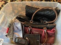 Plastic tote filled with various handbags, small clutches, and makeup bags including a black quilted handbag and silver makeup bag.