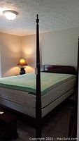Full view of 4 poster bed headboard and mattress including foam pad. Bedpost finial visible. Lamp and room ceiling light in background.