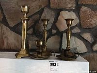 Image showing 5 brass candlesticks arranged on a white shelf in front of a stone wall background, confirming quantity and visible design variety.
