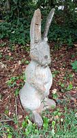 Front and side view of large cement bunny standing upright in a garden bed with leafy background. Damage and cracks visible on left ear and surface dirt.
