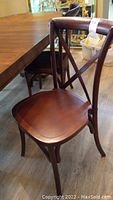 Four solid wood dining chairs with crossed slat backs and curved top rails, brown polished finish. Chairs are shown near a dining table on laminate flooring.