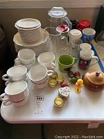 Overview of the lot showing the white ceramic plates stacked, cups arranged, glass pitchers, napkin rings, salt shakers, and wooden bowl on a white table.