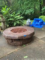 Full side view of round resin fire pit on concrete patio, including propane tank in background and lava rocks inside fire pit center.