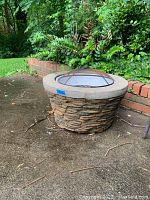Outdoor stone-style fire pit with a concrete top rim, metal grate inside and rusty mesh dome lid, surrounded by garden and brick border.