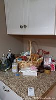 Wicker basket filled with assorted bath and body care products including bottles, jars, soaps, device, and eye mask, displayed on kitchen counter under cabinets.