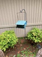 Black metal shepherd hook with a blue metal bird feeder hanging in a garden bed surrounded by green bushes and a beige fence.
