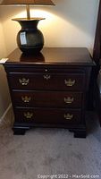 Full front view of dark wood nightstand with lamp on top, showing three drawers with brass handles and the top pull-out drawer closed.