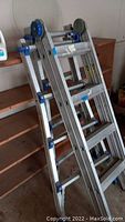 Metal ladder in three sections leaning against a wooden shelving unit in a garage environment, showing overall height and attachments.