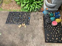 Photo showing black metal wall art, multiple ceramic bird candle holders, plant stand, lanterns, vase, and pot arranged outdoors on concrete with greenery background.