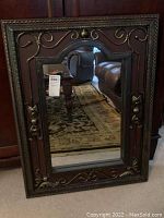 Full view of the large rectangular framed mirror resting against a wall, showing its dark wood frame with decorative metal scrollwork.