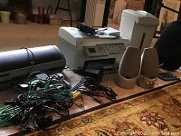 Overall view of printers, speakers, and cables on a carpeted floor showing the HP Photosmart C6180 printer in white, two beige computer speakers, and tangled power and connection cables.