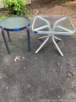 Photo showing two small outdoor side tables on pavement outdoors, round dark framed glass top table and square white framed textured glass top table.