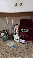 Photo showing burgundy towels, two clear glass candlesticks with white candles, box of Mikasa rose petals, wine goblet, two essential oil bottles, two small sanitizer bottles, and a small decorative bottle.