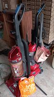 Two upright vacuum cleaners placed side by side in a garage, one Hoover and one Eureka, both red and upright style.