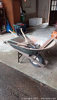Full view of metal wheelbarrow with wood handles seen from front side, including axe, saw, clippers inside.