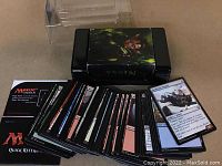 Photo of a black plastic storage box with a textured character illustration on the lid. The box contains approximately 85 Magic the Gathering cards, each standard size with full artwork visible. A Quick Reference guide for the game is also pictured.