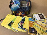 Photo showing over 200 Pokemon cards loosened and spread in front of a collectible metal tin with Charizard and Gyarados images.