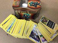 Photo shows over 200 Pokémon trading cards spread out with a Pokémon collectible tin in the background. Cards have yellow borders and various Pokémon characters visible on the front.