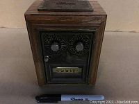 Front view of miniature lockbox, showing oak wood case, metal front panel with two combination dials labeled with letters, lever, and numeric display '5667'. Marker pen placed for scale.