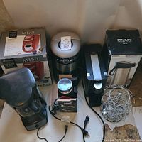 View of the assorted coffee machines, including Keurig Mini, Mr. Coffee drip maker, Stanley air pot, espresso maker, milk frother, and coffee pod carousel on table with visible dust and residue.
