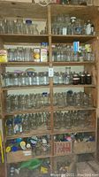 Wide view of wooden shelves filled with multiple rows of various sized clear canning jars, some with blue plastic lids, some open, with boxes and other items on the top shelf and bottoms.