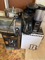 Photo showing all items including the Belgique fry pan on top, Hamilton Beach EasyReach toaster oven beneath, Remington iCoffee steam brew coffee maker in box, Ninja coffee maker, and Moccamaster in box arranged closely together on a floor.