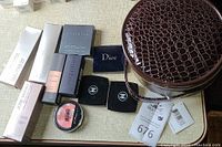 Photo shows multiple boxed cosmetics from brands such as Cover FX, Smashbox, Josie Maran, Laura Mercier, Dior, along with loose Chanel compact powders and Maybelline blush. Cosmetic case visible.