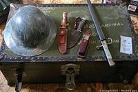 Overall lot showing footlocker lid with helmet, two knives, multi-tool and sword
