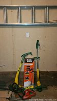 Photo shows the Black & Decker hedge trimmer boxed, green and yellow weed wacker/weed eater, and some 2x6 lumber pieces against a wall under the ladder.