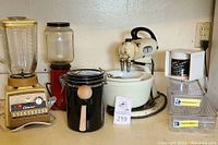 Wide view of the lot showing all four items: the yellow Oster blender on the left, the red KitchenAid coffee grinder next to it, the black canister with wooden scoop in front center, and the old Hamilton Beach stand mixer with bowl and beaters on right.