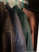 Four men's leather jackets hanging together in a closet from a front angle showing four colors: brown, navy blue, dark green, and tan.