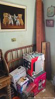 Overview of various boxed appliances stacked against a vintage bed frame and wall with framed cat picture.