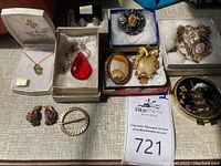 Overview photo of entire lot showing various pendants, earrings, brooches, and a compact on a table surface.