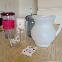 White plastic Kool-Aid Man pitcher with embossed smiling face, handle, and spout alongside Hi-C clear plexiglass mug and other cups on wooden surface.