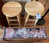 Two natural wood round seat barstools behind a wooden framed, cat-themed tapestry upholstered bench seat with multiple cats depicted on the fabric.
