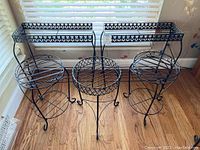 Five black metal plant stands arranged: two rectangular with decorative sides and raised edges, three round lattice-top stands with curved legs.